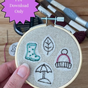 May include: An embroidery hoop with a beige fabric featuring embroidered images of a rain boot, leaf, umbrella, and hat. The text "PDF Download Only" is in a purple starburst. Embroidery floss and needles are in the background.