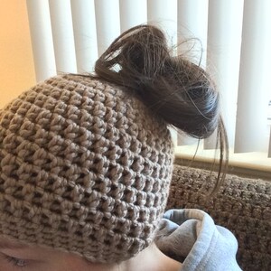 May include: A beige crochet beanie with a textured design. The beanie is worn on the head, with hair pulled through the top. The person is wearing a grey hooded sweatshirt.