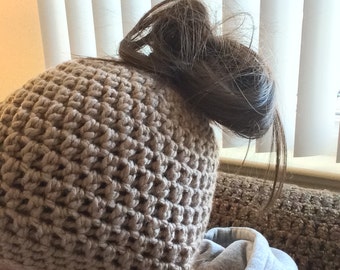 May include: A tan crochet beanie with a textured pattern. The beanie is worn on the head, with hair pulled through the top. The person is wearing a gray hooded sweatshirt.
