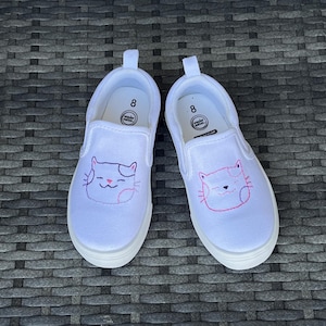 May include: Pair of white slip-on shoes, size 8, with a cat face embroidered on the front. The cat face is outlined in pink and has a smiling mouth. The shoes have a white sole and a pull tab.
