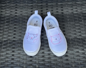 May include: Pair of white slip-on shoes, size 8, with a cat face embroidered on the front. The cat face is outlined in pink and has a smiling mouth. The shoes have a white sole and a pull tab.