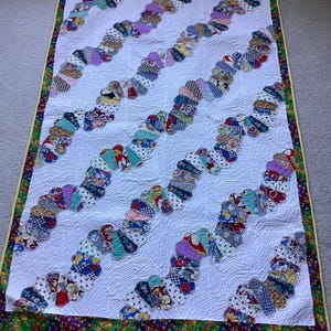 May include: A rectangular quilt featuring a white background with a repeating pattern of diagonal, serpentine rows composed of multi-coloured fabric fan shapes. The quilt is finished with a vibrant, multi-coloured floral border.