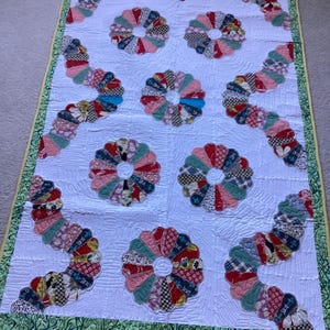 Op de afbeelding: Een rechthoekige quilt met een witte achtergrond en een doorgestikte textuur. Het ontwerp bevat cirkelvormige bloemachtige motieven en kronkelende patronen van veelkleurige lapjes stof. Een groene rand met patroon omlijst de quilt.