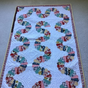 May include: A rectangular quilt featuring a white background with a winding, serpentine pattern made of multi-coloured fabric fan-shaped segments. The border consists of a striped red, white, and black pattern with a thin yellow edge.