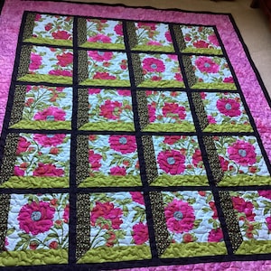 Puede incluir: Un edredón floral con un borde rosa y un patrón de cuadrícula de cuadrados. Cada cuadrado presenta una gran flor rosa sobre un fondo azul claro, enmarcada por acentos verdes y negros. El edredón es probablemente hecho a mano.