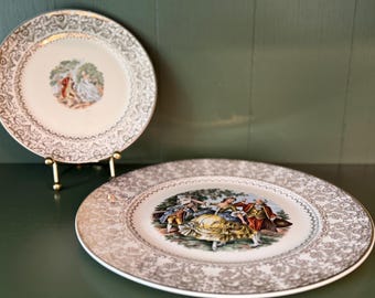 May include: Two round ceramic plates featuring a central illustration of figures in historical attire within a landscape. The rims are decorated with an intricate metallic gold filigree pattern. One plate is displayed upright on a gold metal stand.