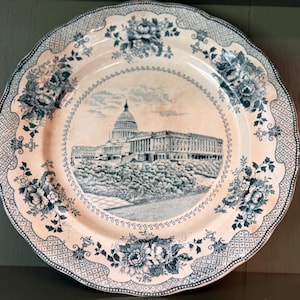 May include: A round ceramic plate featuring a blue and white transferware design. The center depicts the United States Capitol building, while the scalloped rim is decorated with intricate floral patterns and a geometric lattice border.