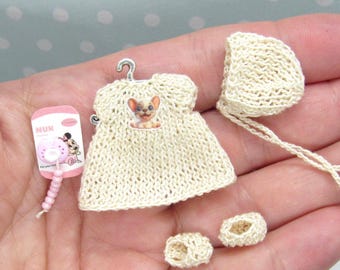 Puede incluir: Una mano sosteniendo un conjunto de ropa de bebé tejida en miniatura, que incluye un vestido, un gorro y botines. El vestido tiene un pequeño diseño de gato en la parte delantera.