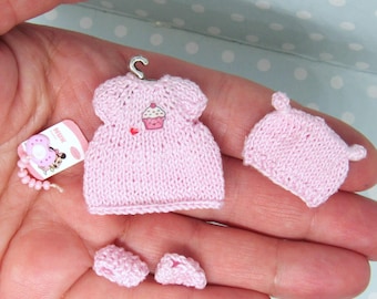 Puede incluir: Un conjunto de bebé de punto rosa con un diseño de cupcake en la camiseta, un gorro a juego y un par de botines.