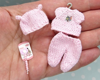 Puede incluir: Un conjunto de bebé de punto rosa con gorro, camisa y pantalón. La camisa tiene un pequeño diseño de gato gris en la parte delantera. Se incluye un sonajero rosa y blanco.