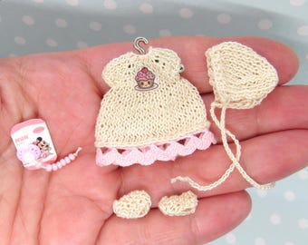 Puede incluir: Un conjunto de ropa de bebé de ganchillo en miniatura, que incluye un vestido, un gorro y botines. El vestido tiene un ribete rosa y un diseño de cupcake. El gorro y los botines son de color crema.