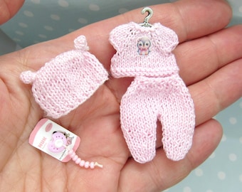 Puede incluir: Un conjunto de bebé de punto rosa con un gorro a juego. El conjunto presenta un pequeño diseño de búho en la parte delantera.