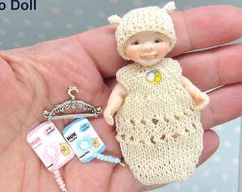 Puede incluir: Un conjunto de muñeca en miniatura con un gorro y un vestido de punto, una percha de plata y dos chupetes. El conjunto es de color crema con un diseño de crochet blanco. Los chupetes son rosa y azul con un diseño de oso y anillo. El texto en los chupetes dice "NUK".
