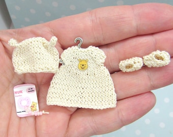 Puede incluir: Un conjunto de ropa de bebé de punto en color crema con un diseño de Winnie de Pooh en el vestido. El conjunto incluye un vestido, un gorro y un par de botines.