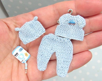 Puede incluir: Conjunto de ropa de bebé en miniatura en azul claro, que incluye un gorro, una camisa con una aplicación de coche y pantalones. También hay un pequeño biberón. El conjunto de punto se muestra en una mano, lo que sugiere una escala pequeña.