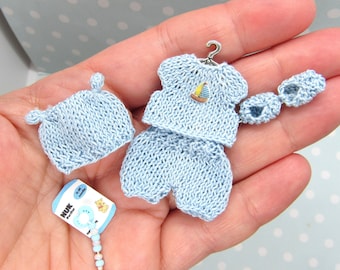 Puede incluir: Un conjunto de bebé de punto azul con un diseño de velero, un gorro a juego y botines. El conjunto se muestra en una mano con un pequeño chupete azul y blanco y una botella de fórmula para bebés.
