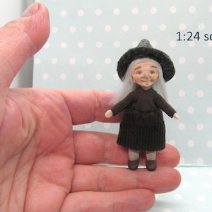 Può includere: Una piccola bambola fatta a mano vestita da strega. Ha i capelli bianchi, un vestito nero e un cappello a punta grigio. La bambola è in scala 1:24.