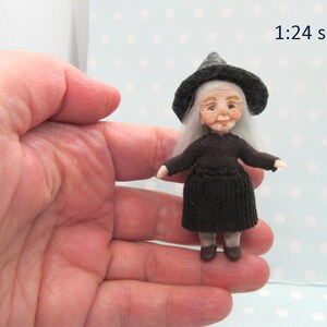 Könnte beinhalten: Eine kleine, handgefertigte Puppe, die als Hexe verkleidet ist. Sie trägt ein schwarzes Kleid und einen schwarzen Hexenhut mit einem grauen Rand. Ihr Haar ist weiß und sie hat ein freundliches Gesicht. Die Puppe ist im Maßstab 1:24.