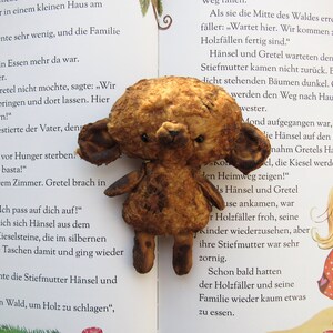 Może przedstawiać: Brązowy miś pluszowy o zużytym, postarzanym wyglądzie siedzi na otwartej książce. Książka jest otwarta na stronie z tekstem w języku niemieckim o historii dwojga dzieci, Hansel i Gretel, kt&oacute;rzy są zagubieni w lesie.