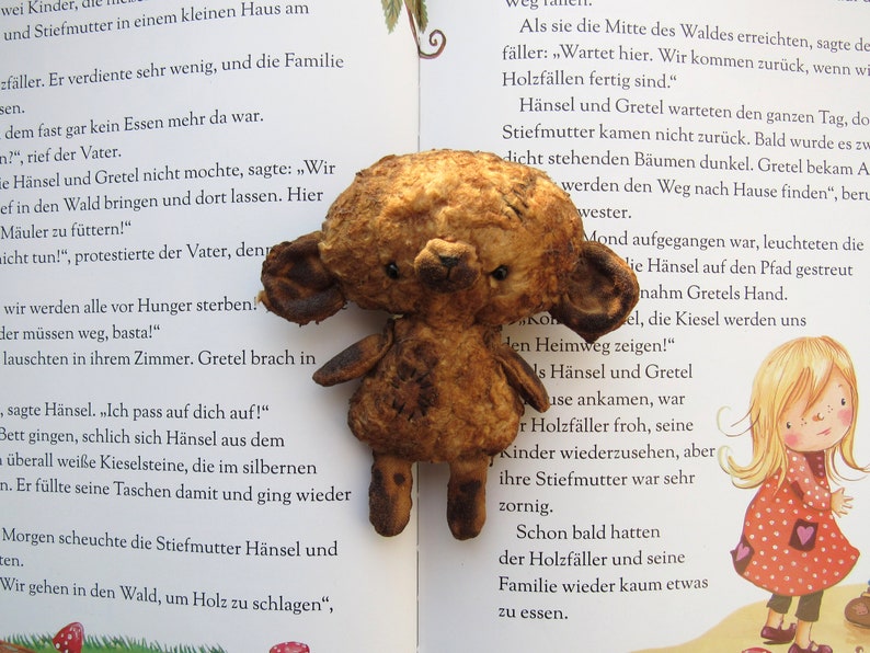 Może przedstawiać: Brązowy miś pluszowy o zużytym, postarzanym wyglądzie siedzi na otwartej książce. Książka jest otwarta na stronie z tekstem w języku niemieckim o historii dwojga dzieci, Hansel i Gretel, kt&oacute;rzy są zagubieni w lesie.