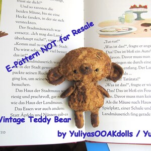 Może przedstawiać: Pluszowy miś vintage z brązowym, zużytym futrem, siedzący na otwartej książce z tekstem w języku niemieckim. Miś ma przyszyty nos i małe czarne oczy. Książka nosi tytuł "Vintage Teddy Bear" i ma niebieskie tło z białym tekstem.
