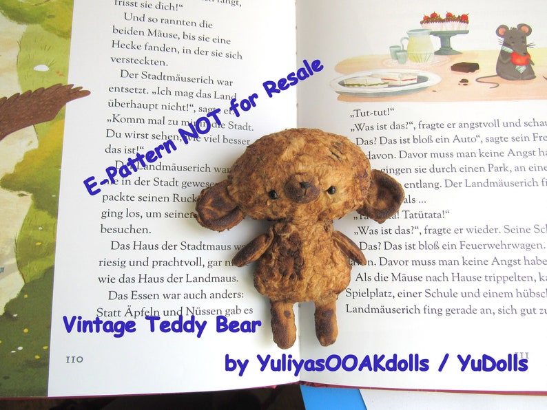 Może przedstawiać: Pluszowy miś vintage z brązowym, zużytym futrem, siedzący na otwartej książce z tekstem w języku niemieckim. Miś ma przyszyty nos i małe czarne oczy. Książka nosi tytuł "Vintage Teddy Bear" i ma niebieskie tło z białym tekstem.