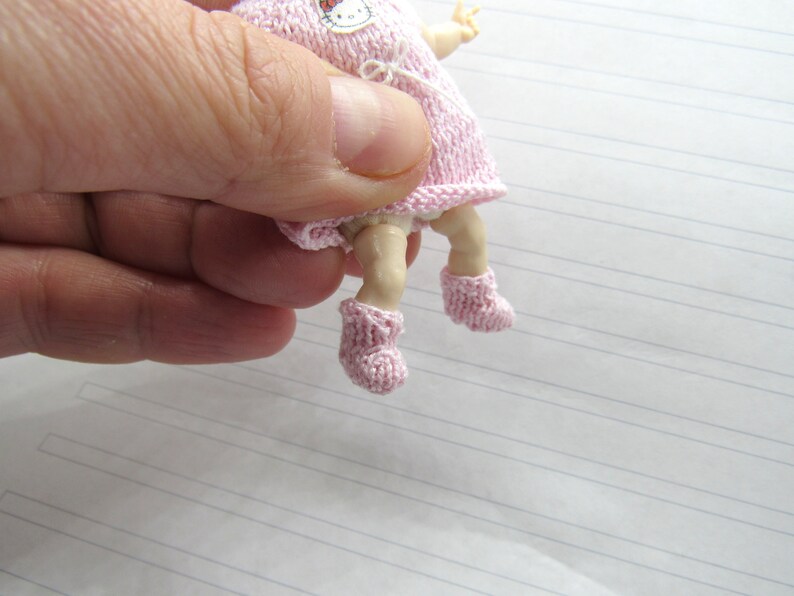 K&ouml;nnte beinhalten: Eine kleine rosa gestrickte Puppe mit einem Hello-Kitty-Motiv auf dem Kleid. Die Puppe tr&auml;gt rosa gestrickte Stiefelchen.