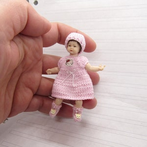 K&ouml;nnte beinhalten: Eine kleine rosa gestrickte Puppe, die eine rosa gestrickte M&uuml;tze mit einer roten Schleife tr&auml;gt. Die Puppe hat ein Hello Kitty-Design auf der Vorderseite ihres Kleides und tr&auml;gt rosa Socken mit einem B&auml;renkopf-Design.