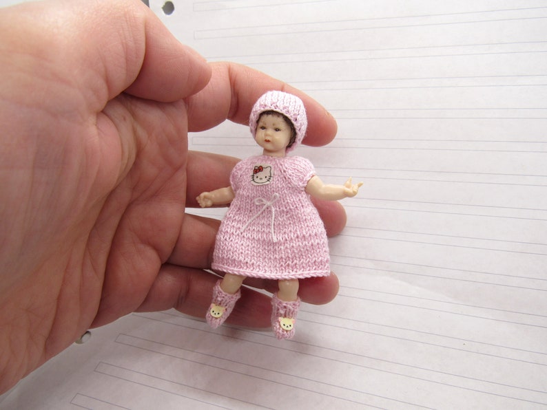 K&ouml;nnte beinhalten: Eine kleine rosa gestrickte Puppe, die eine rosa gestrickte M&uuml;tze mit einer roten Schleife tr&auml;gt. Die Puppe hat ein Hello Kitty-Design auf der Vorderseite ihres Kleides und tr&auml;gt rosa Socken mit einem B&auml;renkopf-Design.