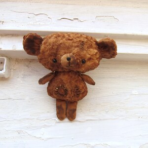 Może przedstawiać: Brązowy miś pluszowy o zużytym, zniszczonym wyglądzie. Miś ma czarne oczy i przyszyty nos. Miś stoi na białym tle.