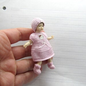 K&ouml;nnte beinhalten: Eine kleine rosa gestrickte Puppe, die eine rosa gestrickte M&uuml;tze und Schuhe tr&auml;gt. Die Puppe hat ein Hello Kitty-Design auf ihrem Kleid.