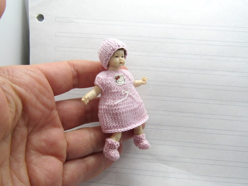 K&ouml;nnte beinhalten: Eine kleine rosa gestrickte Puppe, die eine rosa gestrickte M&uuml;tze und Schuhe tr&auml;gt. Die Puppe hat ein Hello Kitty-Design auf ihrem Kleid.
