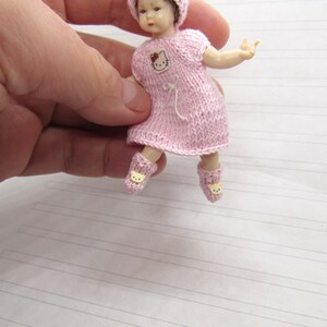 K&ouml;nnte beinhalten: Ein kleines Puppenkleid aus rosa Strick mit einem Hello Kitty-Motiv auf der Vorderseite. Das Kleid wird von einer kleinen Puppe mit rosa Hut und gelben Schuhen getragen.