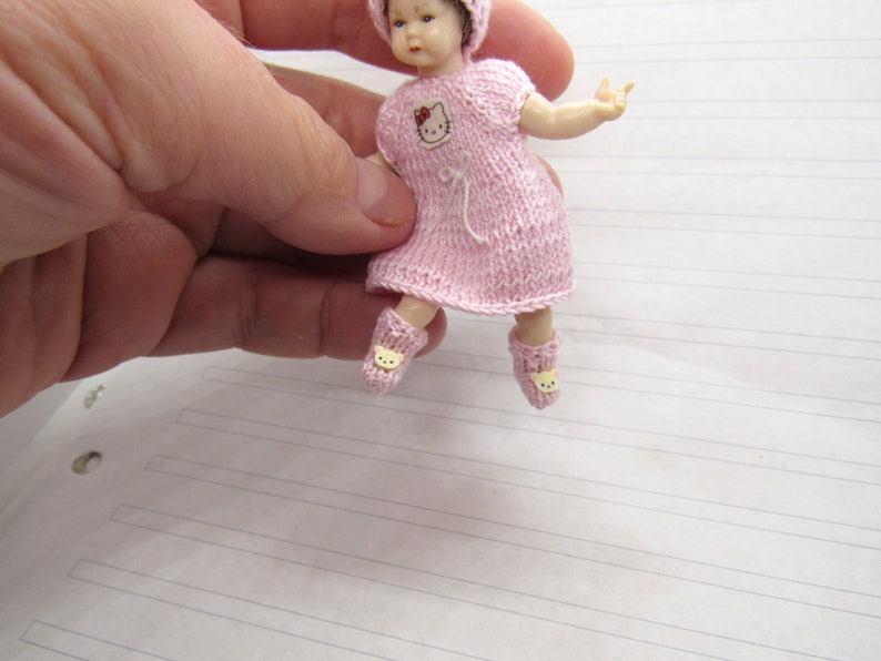 K&ouml;nnte beinhalten: Ein kleines Puppenkleid aus rosa Strick mit einem Hello Kitty-Motiv auf der Vorderseite. Das Kleid wird von einer kleinen Puppe mit rosa Hut und gelben Schuhen getragen.