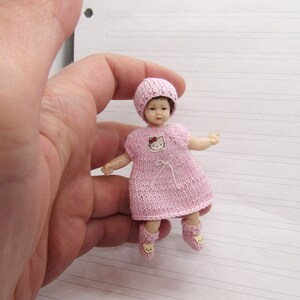 K&ouml;nnte beinhalten: Eine kleine rosa gestrickte Puppe mit einem Hello-Kitty-Design auf dem Kleid. Die Puppe tr&auml;gt eine rosa gestrickte M&uuml;tze und rosa gestrickte Stiefelchen.