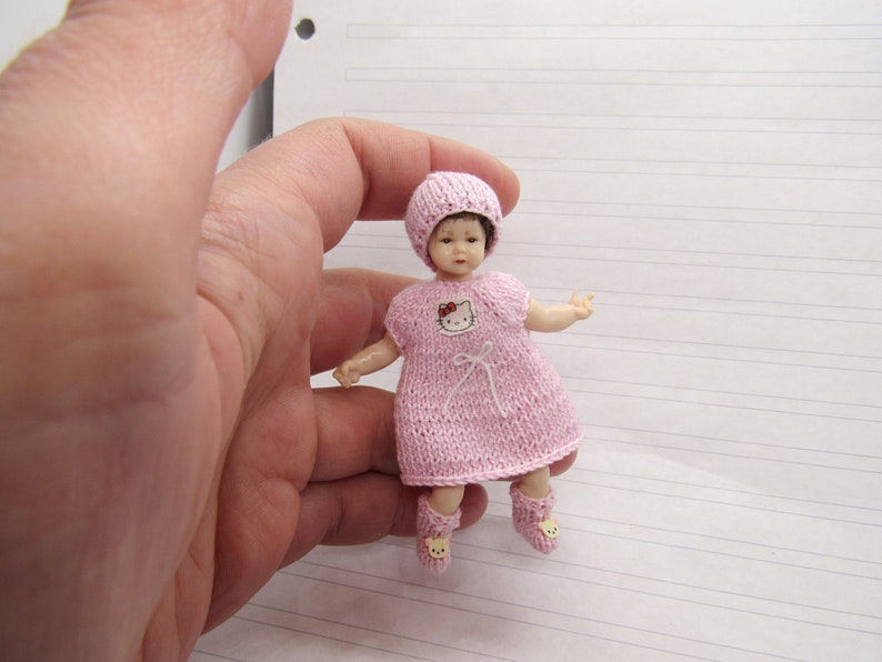 K&ouml;nnte beinhalten: Eine kleine rosa gestrickte Puppe mit einem Hello-Kitty-Design auf dem Kleid. Die Puppe tr&auml;gt eine rosa gestrickte M&uuml;tze und rosa gestrickte Stiefelchen.