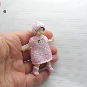 K&ouml;nnte beinhalten: Eine kleine rosa gestrickte Puppe, die eine rosa gestrickte M&uuml;tze mit einer wei&szlig;en Schleife und ein Hello Kitty-Design auf der Vorderseite des Kleides tr&auml;gt. Die Puppe tr&auml;gt rosa gestrickte Stiefelchen.