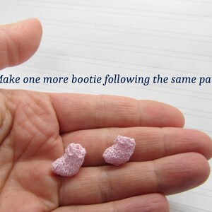 K&ouml;nnte beinhalten: Zwei rosa gestrickte Babyschuhe, einer in jeder Hand. Der Text "Make one more bootie following the same pattern" ist &uuml;ber den H&auml;nden sichtbar.
