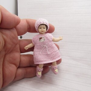 K&ouml;nnte beinhalten: Eine kleine rosa gestrickte Puppe, die ein rosa gestricktes Kleid mit einem Hello-Kitty-Motiv und eine rosa gestrickte M&uuml;tze tr&auml;gt. Die Puppe tr&auml;gt rosa gestrickte Schuhe mit einem Hello-Kitty-Motiv.