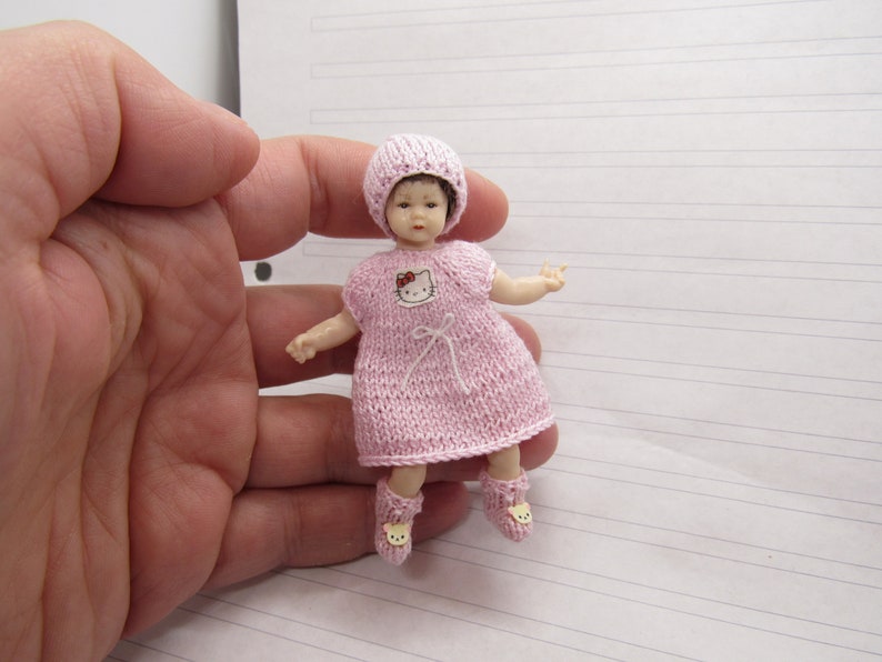 K&ouml;nnte beinhalten: Eine kleine rosa gestrickte Puppe, die ein rosa gestricktes Kleid mit einem Hello-Kitty-Motiv und eine rosa gestrickte M&uuml;tze tr&auml;gt. Die Puppe tr&auml;gt rosa gestrickte Schuhe mit einem Hello-Kitty-Motiv.