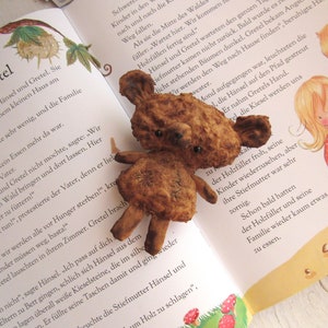 Może przedstawiać: Zbliżenie na brązowego pluszowego misia z czarnymi guzikowymi oczami. Miś siedzi na stronie książki dla dzieci o Jaśku i Małgosi. Książka jest otwarta na stronie z ilustracjami dzieci w lesie.