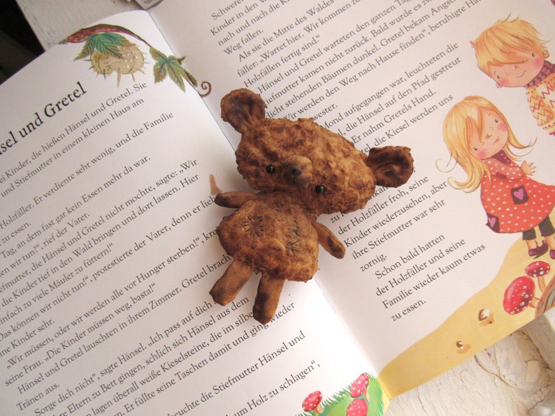 Może przedstawiać: Zbliżenie na brązowego pluszowego misia z czarnymi guzikowymi oczami. Miś siedzi na stronie książki dla dzieci o Jaśku i Małgosi. Książka jest otwarta na stronie z ilustracjami dzieci w lesie.