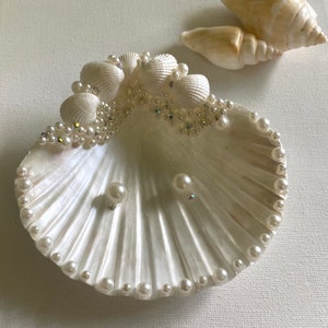 K&ouml;nnte beinhalten: Eine wei&szlig;e Muschelschalen-Schale, die mit Perlen und Strasssteinen verziert ist. Die Schale ist mit zwei Perlenohrringen gef&uuml;llt.
