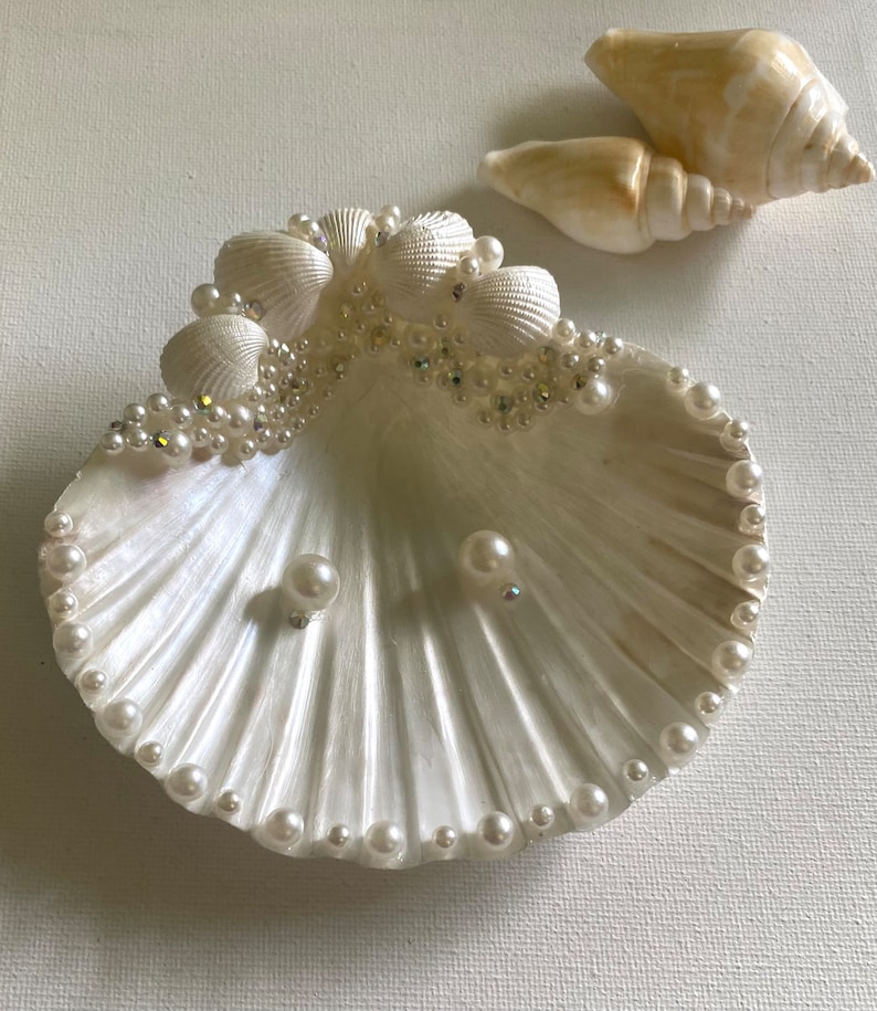 K&ouml;nnte beinhalten: Eine wei&szlig;e Muschelschalen-Schale, die mit Perlen und Strasssteinen verziert ist. Die Schale ist mit zwei Perlenohrringen gef&uuml;llt.