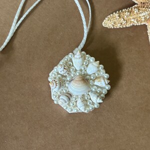 May include: A white hexagon-shaped pendant with a white cord. The pendant is decorated with small white pearls and seashells.
