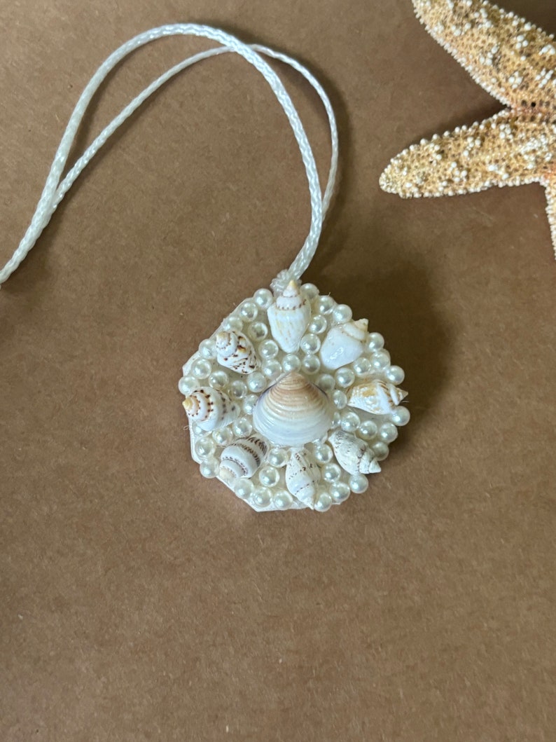 May include: A white hexagon-shaped pendant with a white cord. The pendant is decorated with small white pearls and seashells.