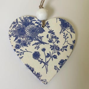 Op de afbeelding: Een hartvormige houten decoratie met een blauw bloemenpatroon op een cr&egrave;mekleurige achtergrond. Het heeft een touwtje om op te hangen en een witte kraal. Het bloemenpatroon bevat verschillende bloemen en bladeren.