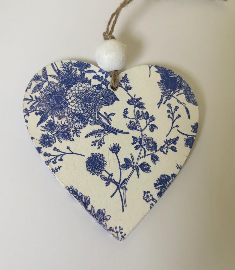 Op de afbeelding: Een hartvormige houten decoratie met een blauw bloemenpatroon op een cr&egrave;mekleurige achtergrond. Het heeft een touwtje om op te hangen en een witte kraal. Het bloemenpatroon bevat verschillende bloemen en bladeren.