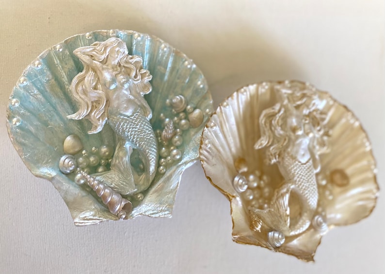K&ouml;nnte beinhalten: Zwei dekorative Muscheln mit einer Meerjungfrauenfigur im Inneren. Die Muscheln sind in Perlmuttwei&szlig; und Hellblau lackiert. Die Meerjungfrauen sind silbern und haben langes flie&szlig;endes Haar.
