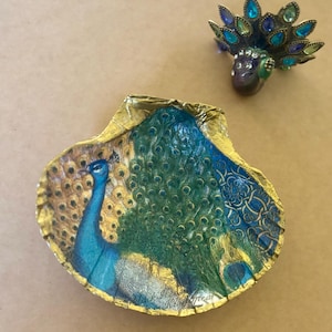 May include: A gold and blue seashell dish with a peacock design. The dish is decorated with gold leaf and has a peacock with a blue tail and a yellow body.